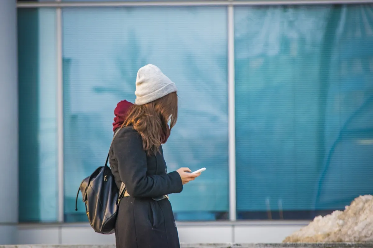 Woman in a knit hat looking at her phone beside a blue glass wall